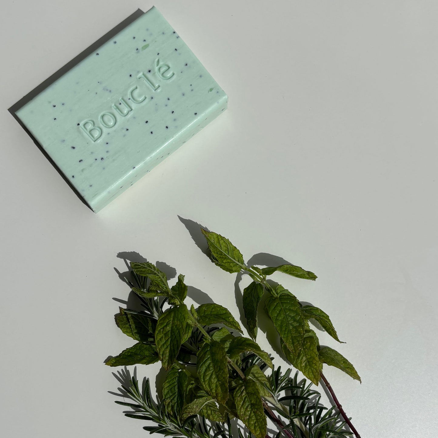 Green soap bar with 'Boucle' branding next to a bundle of herbs on a light gray background