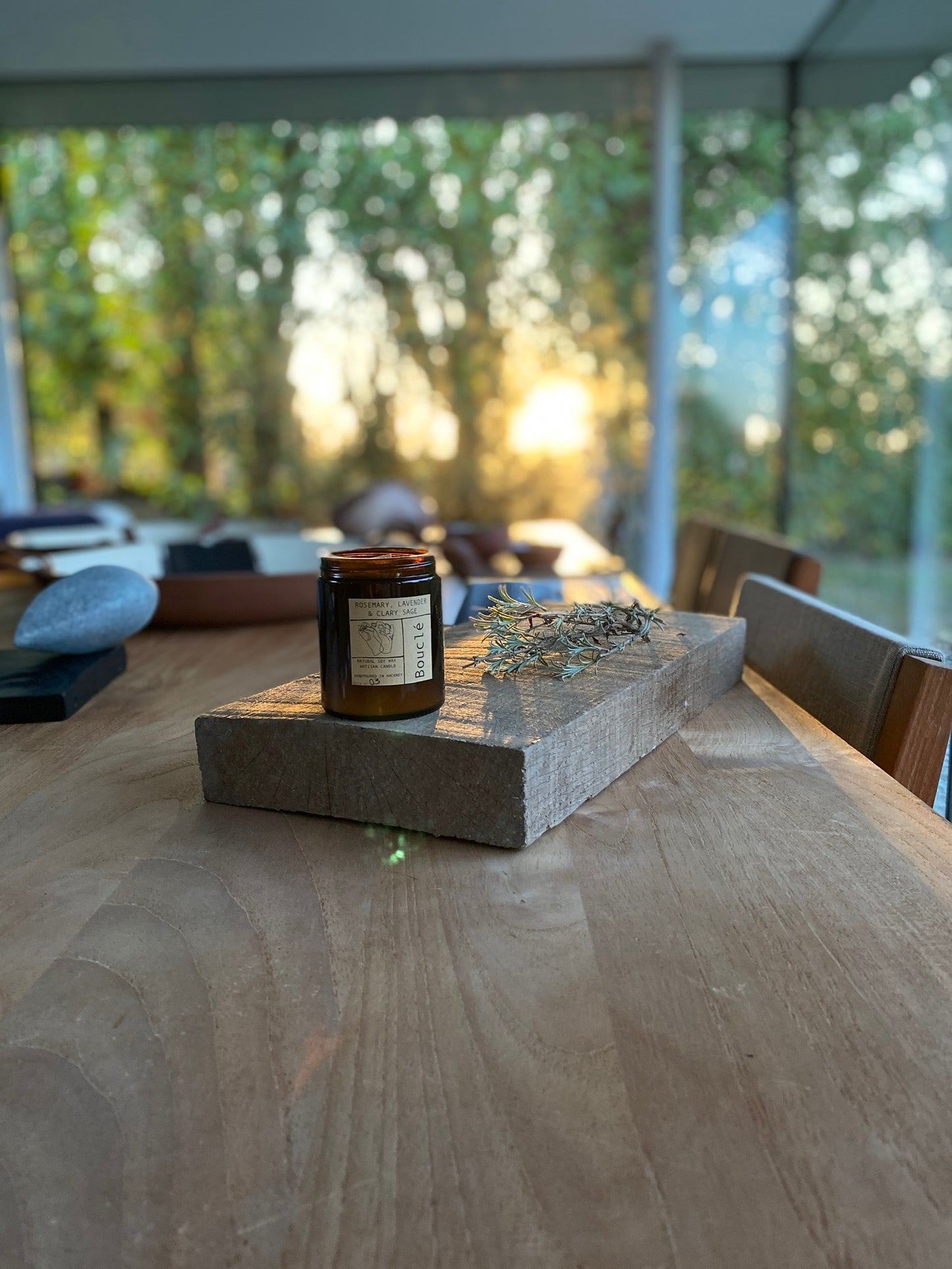 Relaxing Bouclé candle sits on a wooden board as the sun rises behind it. Scented with lavender, rosemary, orange oil and lemon balm it is the perfect scent for summer and all year round.